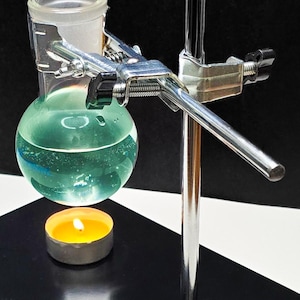 以下が含まれることがあります： 丸底フラスコにターコイズブルーの液体が入っており、金属製のクランプでスタンドに固定されています。点灯したティーライトキャンドルが下にあります。黒い表面に配置されています。