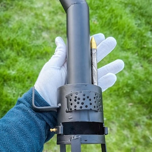 Peut inclure: Un poêle en métal noir avec une cheminée incurvée et un corps cylindrique perforé. Le poêle a un petit raccord en laiton et est soutenu par trois pieds. Une balle décorative est tenue à côté du poêle.