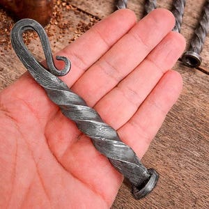 Op de afbeelding: Handgesmede, gedraaide metalen flesopener met een decoratief haakontwerp. De opener heeft een donkergrijze kleur en heeft een textuur, spiraalvormig patroon over de lengte. De basis heeft een cirkelvormig ontwerp.