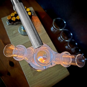 以下が含まれることがあります： 銀色の金属製ハンドルが付いた透明なガラス製の科学機器。 機器には複数の開口部があり、木製の表面に配置されています。 背景には追加のガラスビーカーが見えます。