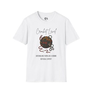 May include: White t-shirt with a graphic featuring a yarn ball with a digital clock, scissors, and the text "Crochet Level." Below the graphic, the text reads "Untangling Yarn Like a Bomb Defusal Expert."