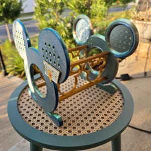 Puede incluir: Un botellero decorativo azul y dorado con forma de orejas de Mickey Mouse. El botellero de madera está sobre una pequeña mesa redonda con una tapa tejida. Las orejas tienen agujeros y una pequeña pieza decorativa.