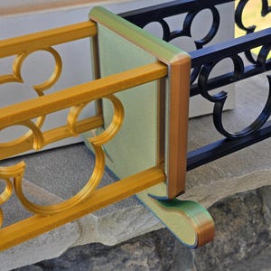 Könnte beinhalten: Ein Abschnitt eines dekorativen Geländers mit einem goldenen Mickey-Mouse-Silhouetten-Design. Das Geländer ist mit einem schwarzen Schmiedeeisenzaun verbunden. Das Eckstück hat eine grüne und braune Verkleidung, und das Geländer steht auf einer Steinoberfläche.