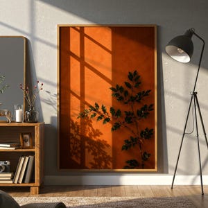 May include: A large rectangular wall art piece in a wooden frame featuring a deep orange background with a dark silhouette of a leafy plant branch and a window pane shadow cast across the surface, displayed on a wall next to a black tripod floor lamp.
