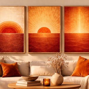 May include: Three framed wall art pieces depicting a sunset. Each artwork has a gradient of orange and red tones, with a sun or sunburst design. The art hangs above a cream sofa with orange cushions.