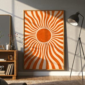 Puede incluir: Un cuadro enmarcado con un diseño retro de rayos de sol en naranja y crema. La obra de arte se exhibe en una habitación con una estantería de madera, un espejo y una lámpara de pie negra.