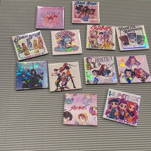 May include: A collection of square, holographic-effect pinback buttons with various colorful designs. The buttons feature illustrations of characters, text, and graphics. Some buttons have text that reads "Sata Boys", "My Little Soda Pop", and "Huntrix Ramyeon".