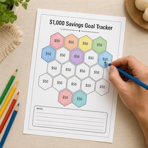 May include: A white paper sheet titled $1,000 Savings Goal Tracker featuring a honeycomb pattern of hexagons, each labeled $50. Some hexagons are colored in pink, orange, yellow, green, blue, and purple, with a notes section at the bottom.