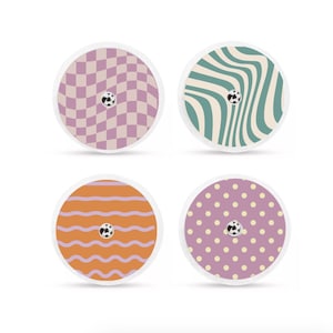 Op de afbeelding: Vier ronde decoratieve stickers met verschillende patronen. Eén heeft een paars en wit dambordpatroon, een andere groene en crèmekleurige golvende lijnen, een derde oranje horizontale lijnen en de laatste paarse stippen. Elk heeft een klein zwart-wit midden.