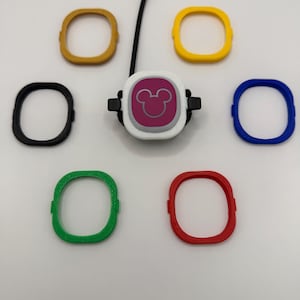 May include: A white button with a pink Mickey Mouse icon and a black cord, surrounded by six colorful plastic rings in yellow, gold, black, blue, green, and red.