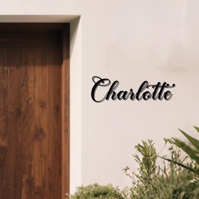 May include: Black metal sign with the name "Charlotte" in a cursive font. The sign is mounted on a white wall next to a wooden door and greenery.