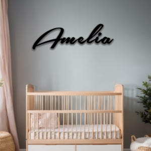 May include: Black cursive metal sign that reads "Amelia" mounted on a light blue wall above a wooden crib. The crib has a white mattress and a pink pillow. A pink curtain is on the left.