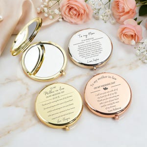 May include: Three round metal compact mirrors in gold and rose gold finishes. One mirror is open, revealing a reflective surface. The other two are closed, displaying engraved text addressed to a mother and mother-in-law for a wedding day.