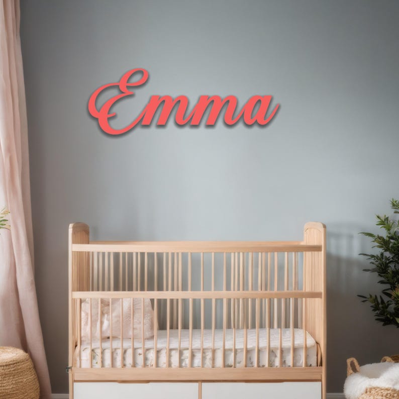 May include: Coral-colored wooden sign spelling the name "Emma" in a cursive font, mounted on a light blue wall. A wooden crib is below the sign, with a white mattress and a pink pillow. A plant and a woven basket are in the background.