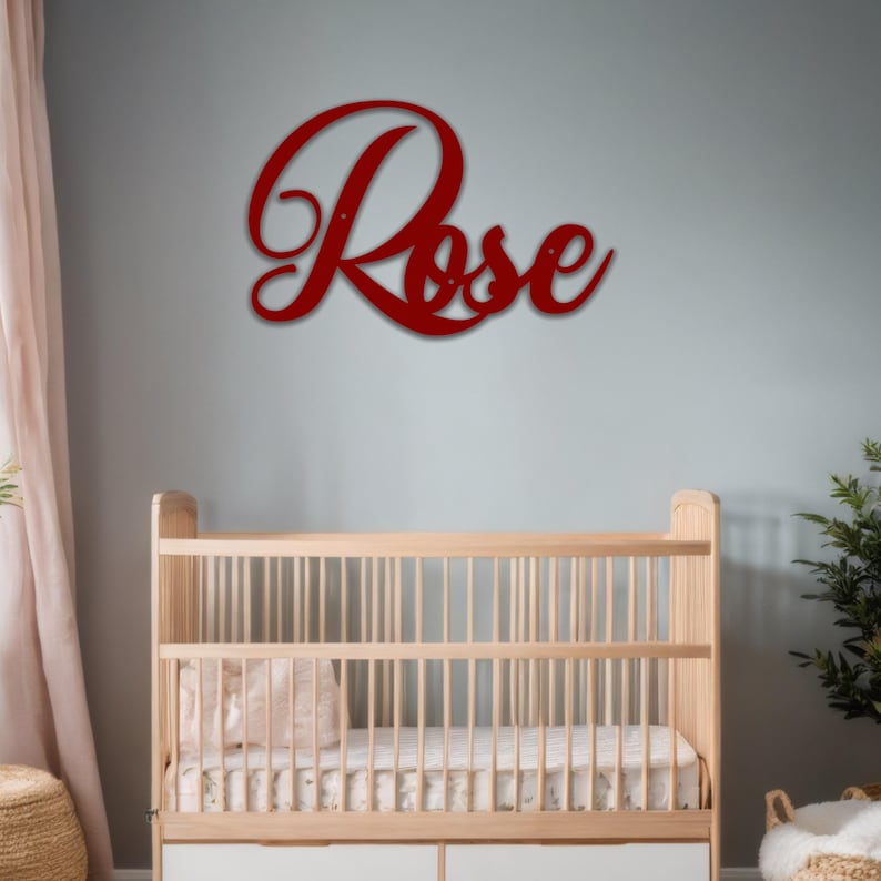 May include: A red metal sign that spells out the name "Rose" in a cursive font. The sign is mounted on a light gray wall above a wooden crib. The crib has a white mattress and a light pink blanket.