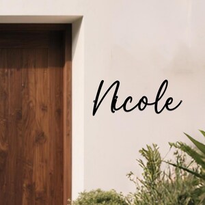 May include: Black metal sign with the name "Nicole" in a cursive font. The sign is mounted on a white wall next to a wooden door and some green plants.