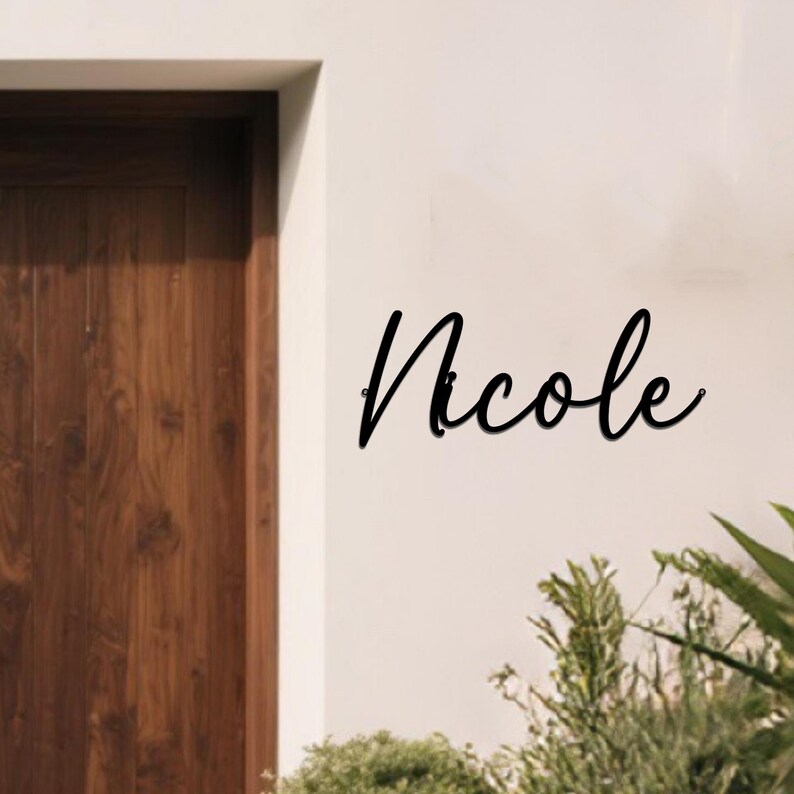 May include: Black metal sign with the name "Nicole" in a cursive font. The sign is mounted on a white wall next to a wooden door and some green plants.