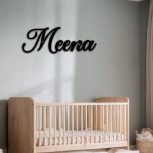 May include: Black cursive wooden sign that spells out the name "Meena". The sign is mounted on a light green wall above a wooden crib. The nursery has a neutral color palette.