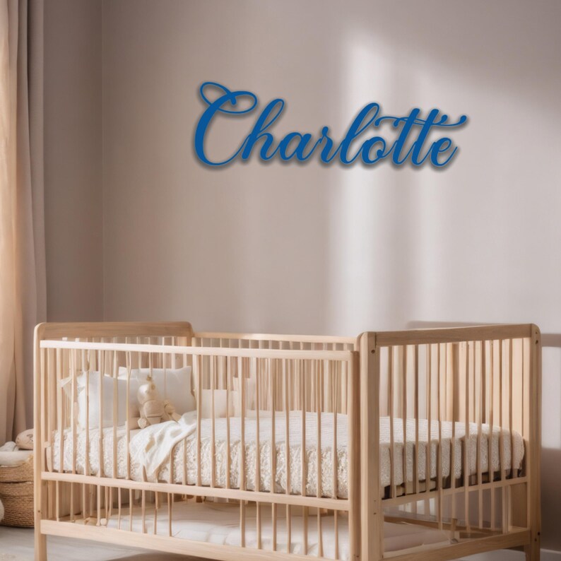 May include: A blue cursive name "Charlotte" is mounted on a wall above a wooden crib. The crib is filled with white pillows, a blanket, and a stuffed animal. The room has a neutral color palette.