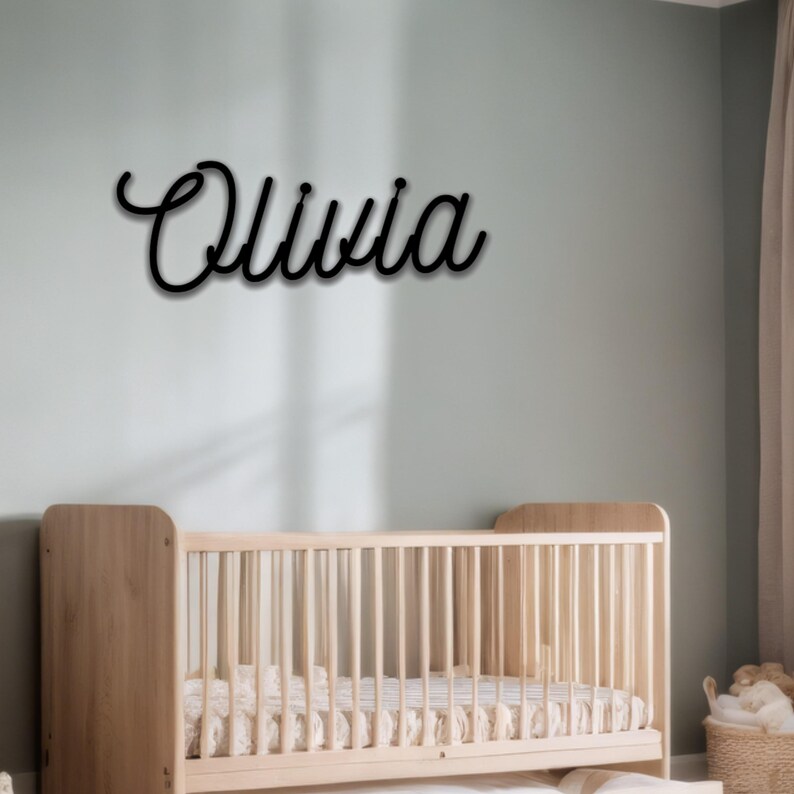 May include: Black cursive wall art spelling the name "Olivia". The name is mounted on a light green wall above a wooden crib. The nursery decor includes a woven basket and a sheer curtain.