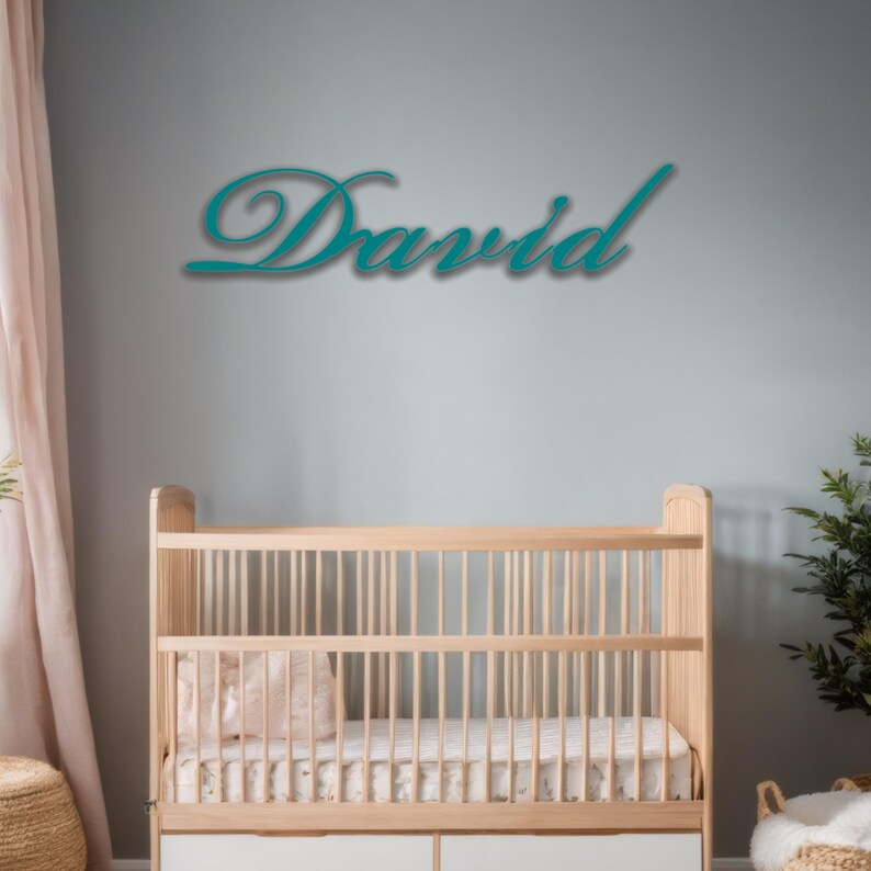 May include: A teal-colored wooden sign that spells out the name "David" in a cursive font. The sign is mounted on a light gray wall above a wooden crib with a white mattress and a pink pillow.