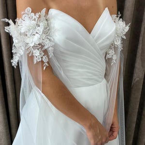 Peut inclure: Une robe de mariée blanche avec un décolleté en cœur et un corsage drapé. La robe présente de délicates dentelles et des ornements floraux sur les épaules, avec un tissu transparent formant des manches. La robe est faite d'un tissu doux et fluide.