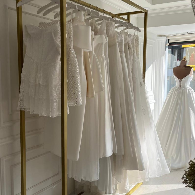 Puede incluir: Exhibici&oacute;n de vestidos de novia blancos colgados en una estructura dorada. Los vestidos var&iacute;an en estilo, incluyendo encaje, sat&eacute;n y dise&ntilde;os sencillos. Un maniqu&iacute; con un vestido de novia es visible en el fondo.