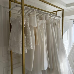 Puede incluir: Una colecci&oacute;n de vestidos de novia blancos colgados en una barra de metal dorado. Los vestidos var&iacute;an en estilo, incluyendo dise&ntilde;os de encaje, lentejuelas y sat&eacute;n. La barra est&aacute; en una habitaci&oacute;n luminosa con paredes blancas.