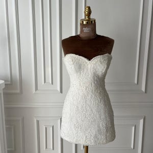 Puede incluir: Un minivestido blanco sin tirantes con intrincados detalles de encaje. El vestido se exhibe en un maniqu&iacute; marr&oacute;n con una parte superior dorada. El maniqu&iacute; tiene las palabras "madame amour" impresas.