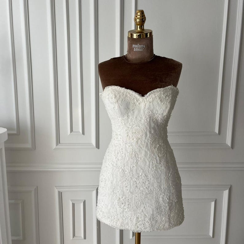 Puede incluir: Un minivestido blanco sin tirantes con intrincados detalles de encaje. El vestido se exhibe en un maniqu&iacute; marr&oacute;n con una parte superior dorada. El maniqu&iacute; tiene las palabras "madame amour" impresas.