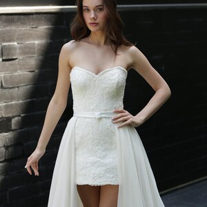 Puede incluir: Un vestido de novia blanco sin tirantes con escote coraz&oacute;n y falda corta, cubierto por una falda fluida y transparente. El corpi&ntilde;o est&aacute; adornado con intrincados detalles de encaje, y un cintur&oacute;n blanco fino ci&ntilde;e la cintura. El vestido se usa frente a una pared de ladrillos oscuros.