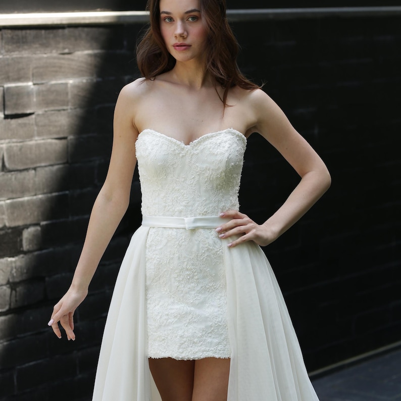 Puede incluir: Un vestido de novia blanco sin tirantes con escote coraz&oacute;n y falda corta, cubierto por una falda fluida y transparente. El corpi&ntilde;o est&aacute; adornado con intrincados detalles de encaje, y un cintur&oacute;n blanco fino ci&ntilde;e la cintura. El vestido se usa frente a una pared de ladrillos oscuros.