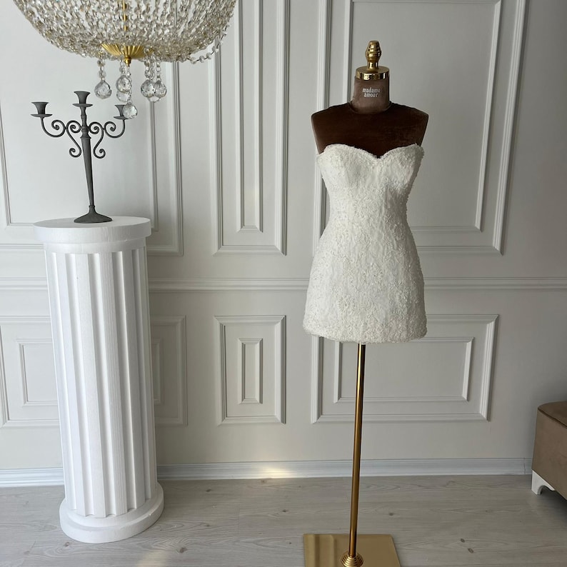 Puede incluir: Un minivestido blanco sin tirantes sobre un maniqu&iacute;. El vestido tiene un aspecto texturizado, similar al encaje. El maniqu&iacute; es marr&oacute;n en la parte superior y dorado en la base. Un pilar blanco y un candelabro negro est&aacute;n en el fondo.