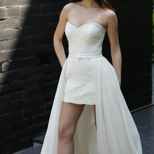 Puede incluir: Vestido de novia blanco sin tirantes con escote coraz&oacute;n y corpi&ntilde;o corto y ajustado. El vestido presenta intrincados detalles de encaje y una sobrefalda fluida y transparente. Un cintur&oacute;n blanco fino ci&ntilde;e la cintura. Para una ocasi&oacute;n formal.