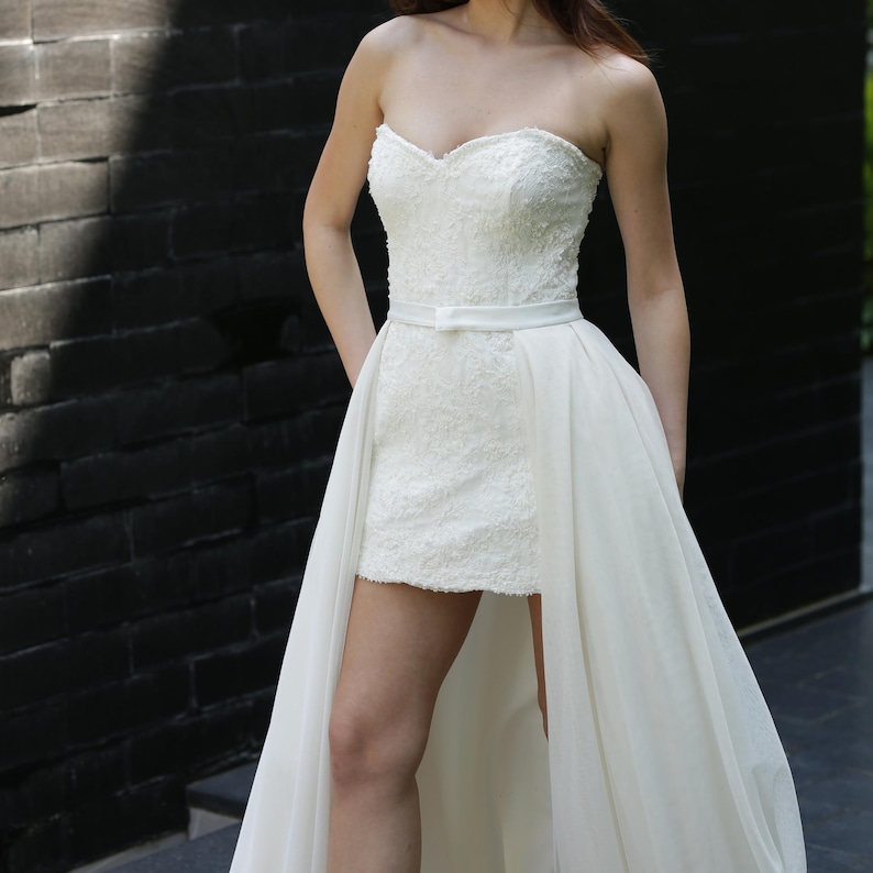 Puede incluir: Vestido de novia blanco sin tirantes con escote coraz&oacute;n y corpi&ntilde;o corto y ajustado. El vestido presenta intrincados detalles de encaje y una sobrefalda fluida y transparente. Un cintur&oacute;n blanco fino ci&ntilde;e la cintura. Para una ocasi&oacute;n formal.