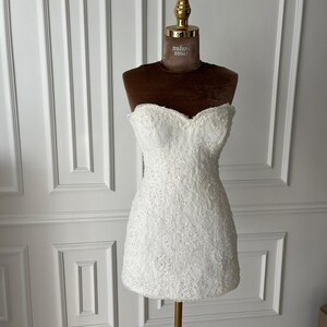 Puede incluir: Un minivestido blanco sin tirantes con intrincados detalles de encaje. El vestido se exhibe en un maniqu&iacute; con un busto de terciopelo marr&oacute;n y un cuello dorado. El vestido tiene un escote coraz&oacute;n y una silueta ajustada.