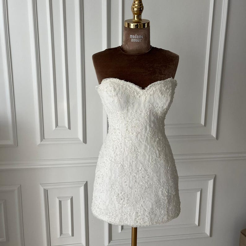 Puede incluir: Un minivestido blanco sin tirantes con intrincados detalles de encaje. El vestido se exhibe en un maniqu&iacute; con un busto de terciopelo marr&oacute;n y un cuello dorado. El vestido tiene un escote coraz&oacute;n y una silueta ajustada.