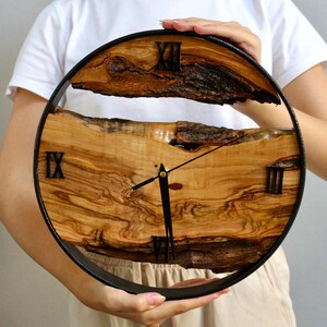 May include: A round wooden clock with a black metal frame. The clock face is made of natural wood with visible grain and texture. Black Roman numerals mark the hours. The clock hands are black.