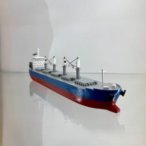 Pode incluir: Modelo de navio de carga com casco vermelho, seção média azul e convés cinza. O modelo apresenta superestruturas brancas, guindastes e um mastro. O navio é uma réplica detalhada de um navio comercial.