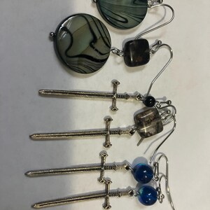 Peut inclure: Une collection de boucles d'oreilles faites à la main. Les boucles d'oreilles présentent divers modèles, notamment des perles ovales marbrées vertes et noires, des perles carrées marron, des petites perles noires et des breloques épées argentées. Certaines boucles d'oreilles ont des perles de verre bleues.