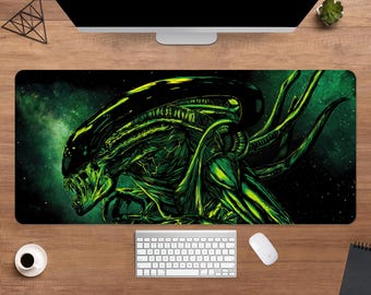 May include: A rectangular mouse pad featuring a detailed illustration of an Alien creature in shades of green and black against a dark green and black space-themed background. The mouse pad is shown with a keyboard, mouse, and coffee cup.