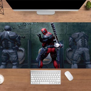 May include: A rectangular desk mat featuring a comic book scene with Deadpool in a red and black suit, holding a weapon. Two figures in gray armor are in the background. The words "the END" are visible on the wall. A keyboard and mouse are in the foreground.