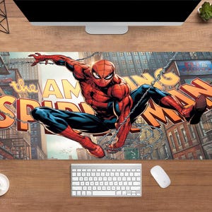 May include: A rectangular desk mat featuring the Amazing Spider-Man in a dynamic pose. The mat displays a comic book illustration of Spider-Man in a red and blue suit, with the words "the AMAZING SPIDER-MAN" in large, orange letters.