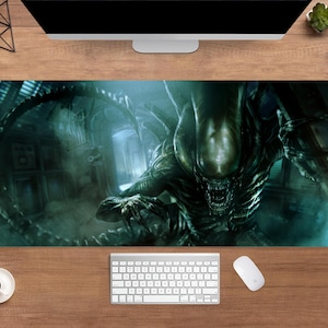 May include: A large rectangular desk mat featuring a detailed image of an Alien creature in a dark, industrial setting. The creature is predominantly dark green with sharp teeth and claws. The background is a mix of blues and greens.