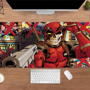 May include: A rectangular desk mat featuring a comic book style illustration of Deadpool with a skull face, holding a gun. The mat has a brick wall background with comic book panels and red "X" marks. A keyboard, mouse, and coffee cup are visible.