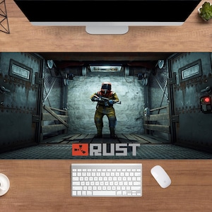 May include: A large rectangular desk mat featuring a scene from the video game "Rust." The image shows a figure in a hazmat suit holding a weapon inside a metal structure. The word "RUST" is displayed in orange and white text at the bottom.