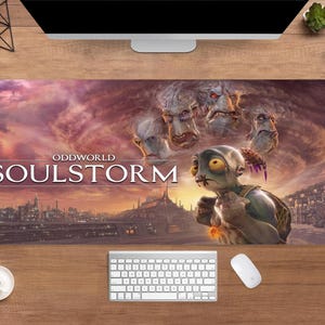May include: A large rectangular mousepad featuring the Oddworld: Soulstorm video game. The design includes the game's title, a character with large yellow eyes, and several grotesque faces against a cloudy, orange sky. A keyboard and mouse are visible.