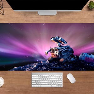 Puede incluir: Alfombrilla de escritorio rectangular con el personaje animado WALL-E sobre un fondo estrellado púrpura y rosa. El robot está representado sobre una superficie rocosa. Se ven un teclado, un ratón y una taza de café.