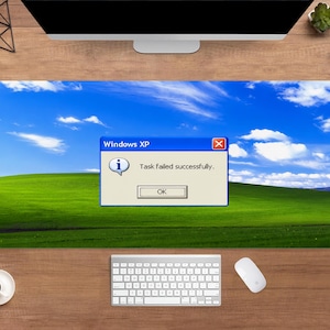 May include: A rectangular desk mat featuring the iconic Windows XP "Bliss" wallpaper with a blue sky, green hills, and a pop-up window that reads "Task failed successfully." A keyboard, mouse, and coffee cup are also visible.