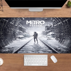 May include: A rectangular black and white desk mat featuring the game title "Metro Exodus." The design depicts a post-apocalyptic scene with a figure walking on train tracks. A keyboard, mouse, and coffee cup are visible.